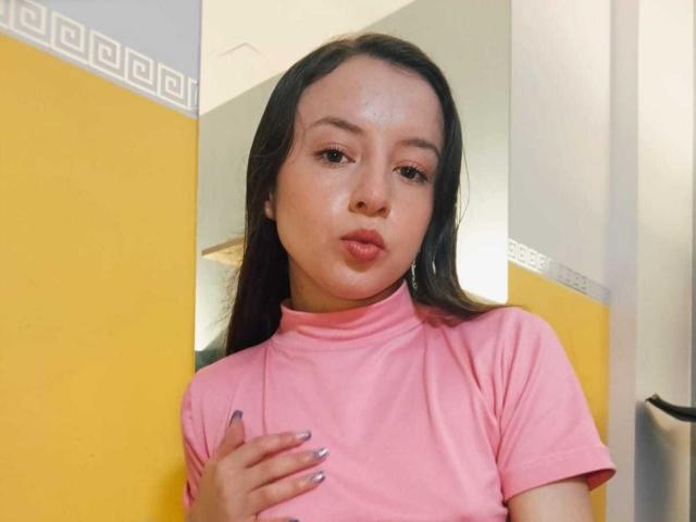 VioletaNova, puffy pink t-shirt, hand on chest, silver nail polish, pouty lips, long brown hair, yellow and white wall, mirror reflection, soft lighting, subtle makeup, cute.