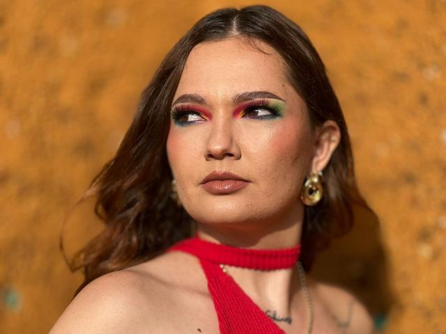 Close-up of HecateCorvin with brown wavy hair, wearing a red knit halter top. She has bold makeup with green and pink eyeshadow, red eyeshadow, and dark eyeliner. She's looking to the side with a serious expression. Gold earrings are visible. Skin is fair with a slight sheen. The background is a textured, orange-brown surface. Light and shadow emphasize her facial features. Her makeup and earrings are the focal points.