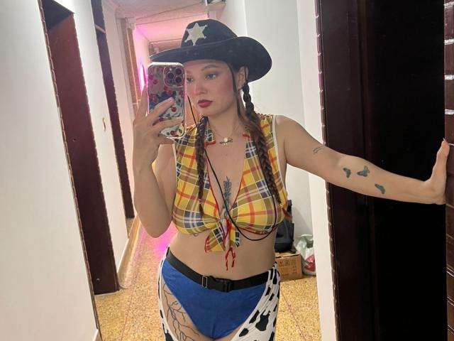 HecateCorvin with small tits, wearing a cowboy hat, plaid crop top, blue and cowprint shorts, holding phone in mirror selfie, showing tattoos on arm and thigh.