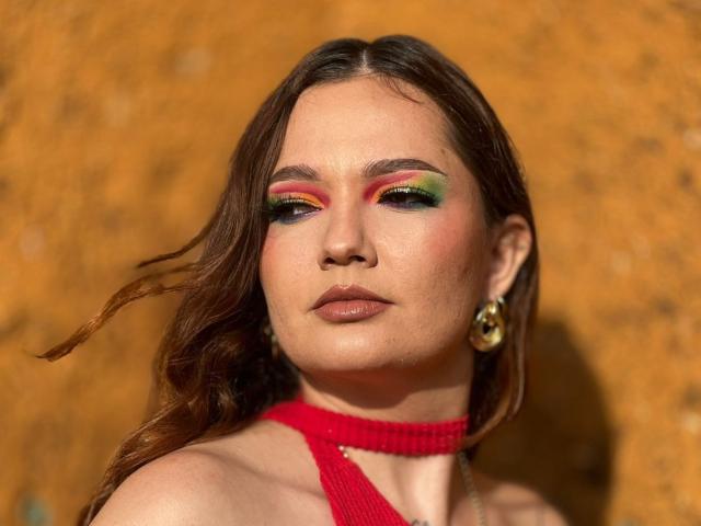 HecateCorvin with colorful eyeshadow, red halter top, and gold earrings, has brown wavy hair, and a serious expression, showing her slightly flushed, makeup-covered face.