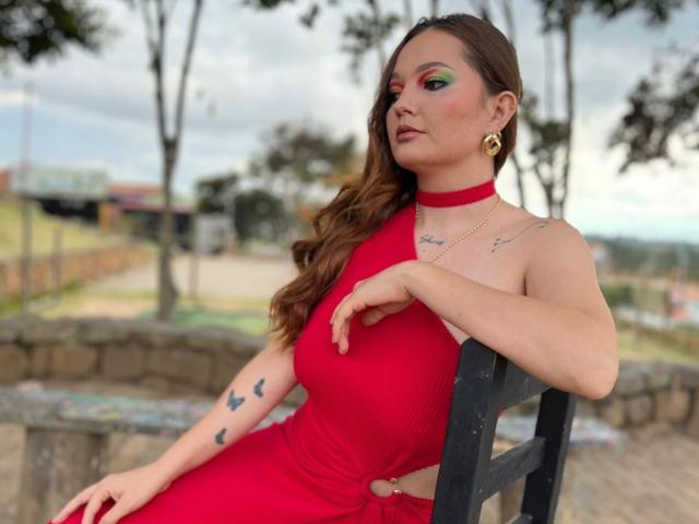 HecateCorvin sits on a black chair, wearing a bright red, sleeveless dress with a cutout side. She has long, wavy brown hair and is looking to the right. Her makeup is bold with green eyeshadow. She has gold hoop earrings and small tattoos on her left arm and right shoulder. Her right arm rests on the chair's back. The image is a photograph with a slightly blurred background of trees and a stone wall. The overall composition is balanced with HecateCorvin as the central focus.