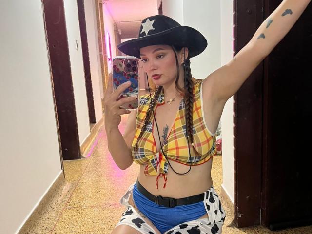 HecateCorvin with braided hair, cowboy hat, yellow plaid top, showing tits, blue cow print shorts, taking a mirror selfie, arm tattoo, holding phone with pink case.