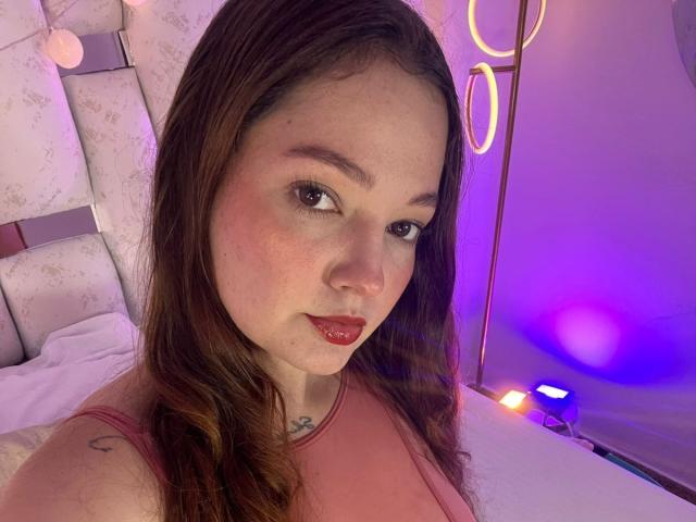 HecateCorvin has long brown hair, red lipstick, freckles, and a small crescent moon tattoo on her shoulder. She's wearing a pink top, looking at the camera.