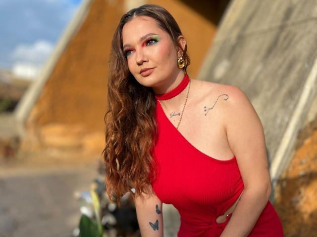 HecateCorvin with long brown wavy hair, red one-shoulder dress, green and pink eye makeup, gold earrings, butterfly tattoos, and a red choker, leaning on a motorcycle.