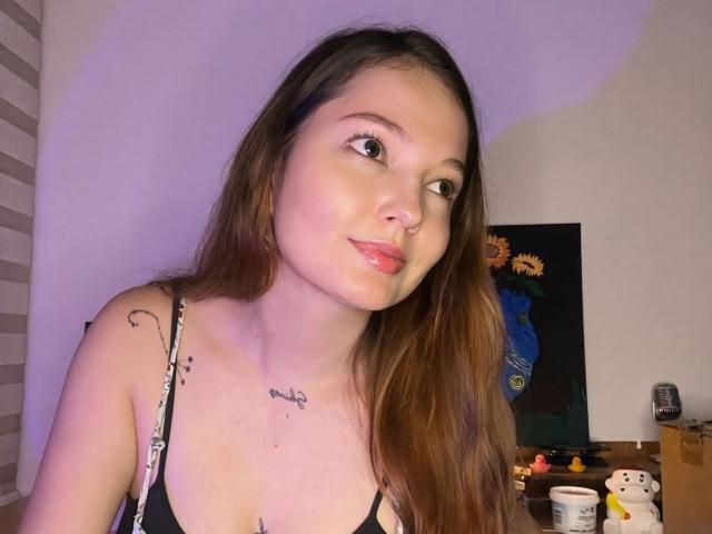 HecateCorvin with long brown hair, looking to the side, pink lips, small tattoos on shoulder and chest, wearing black spaghetti strap top, showing her tits.