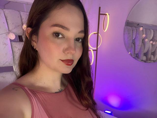 HecateCorvin, a young woman with fair skin and long brown hair, stares directly at the camera with a slight smile. She has red lipstick, subtle eye makeup, and a small tattoo on her right shoulder. She wears a pink sleeveless top. The background includes a neon light and a circular mirror reflecting abstract shapes. The purple and blue lighting adds a modern, slightly moody atmosphere. Her small ear stud is visible on her left ear.