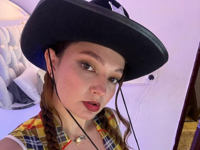 HecateCorvin with braided brown hair, wearing a black hat, plaid shirt, and red lipstick, has earbuds in, with a subtle tattoo on her chest, and a slightly pouty expression.