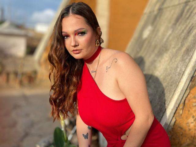HecateCorvin with wavy brown hair, red halter dress, gold hoop earrings, and tattoos, leaning against a stone wall, showing her tits and looking at the camera.