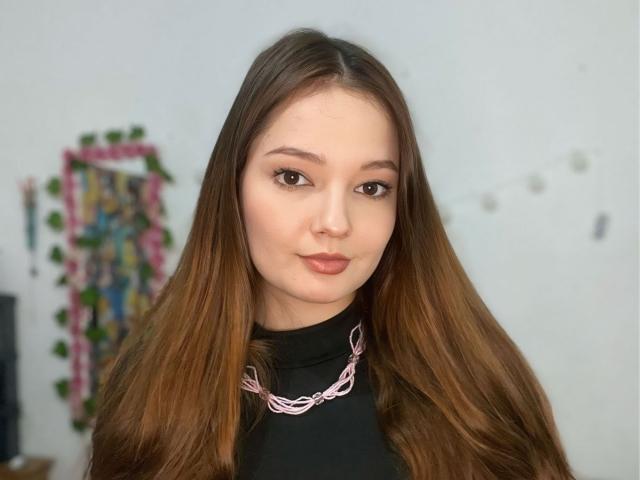 HecateCorvin has long brown hair, light skin, and full lips with subtle pink lipstick. She wears a black turtleneck and a pink chain necklace, looking directly at the camera.