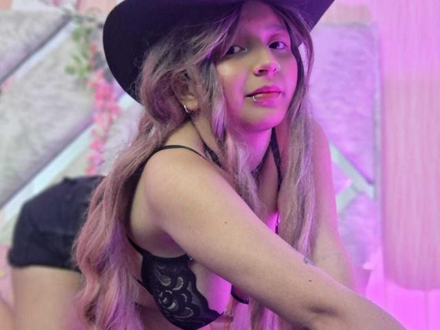 CandySwan with long pinkish-blonde hair, black hat, black lace bra, and black shorts, leaning forward, showing small tits and a nose ring, under purple lighting.