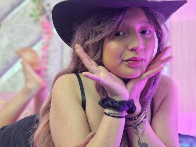 CandySwan with pinkish-purple hair, black hat, and black tank top, pouting with hands on cheeks, showing a lip ring and black scrunchie on wrist, laying on bed.