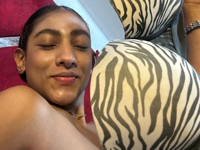 NatalyBlonder with brown skin, closed eyes, and a slight smile, sticking out her tongue. Her bare shoulders and zebra-patterned ass are in the foreground. She's wearing a necklace.