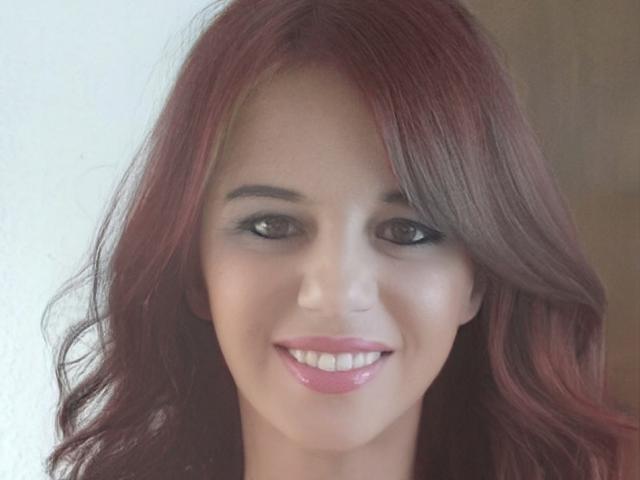 Close-up of CendySander with light skin and reddish-brown wavy hair. She has dark brown eyes with subtle eyeliner and full pink glossy lips. Her eyebrows are thin and well-groomed. CendySander's hair has a slight wave, falling to the sides of her face. The image is centered on her face, capturing her cheerful expression. The background is a simple, light-colored wall. The texture of her hair is smooth and slightly shiny. The overall composition focuses on her facial features and expression.