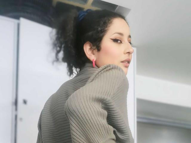 IrinaSweett with curly black hair, pink earrings, and a ribbed gray sweater, looking over her shoulder with a slight smile. Her makeup is subtle with winged eyeliner.