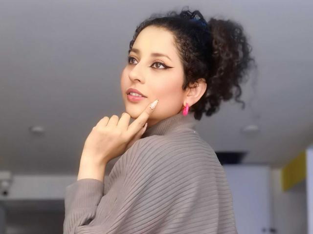 IrinaSweett with curly black hair in a ponytail, wearing a grey ribbed sweater, pink earrings, and touching her lip with a finger, has a cute smile and winged eyeliner.