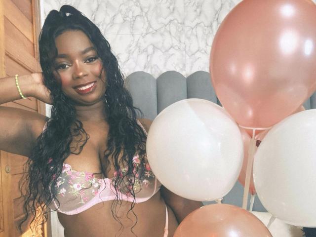 SamyLorenss with dark skin, long curly black hair, smiling, wearing pink floral bra, holding pink and white balloons, wood door on left, marble wallpaper in background.