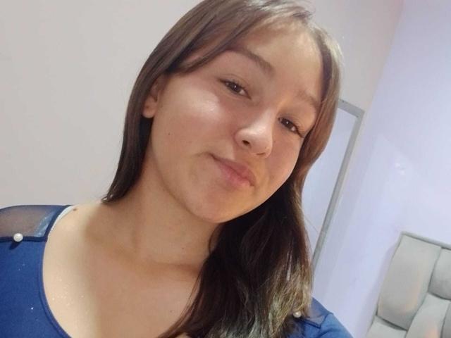EmmaQuintana with light brown hair, slightly wavy, wearing a blue top, has a subtle smile, brown eyes, and a clear complexion, in a casual selfie.