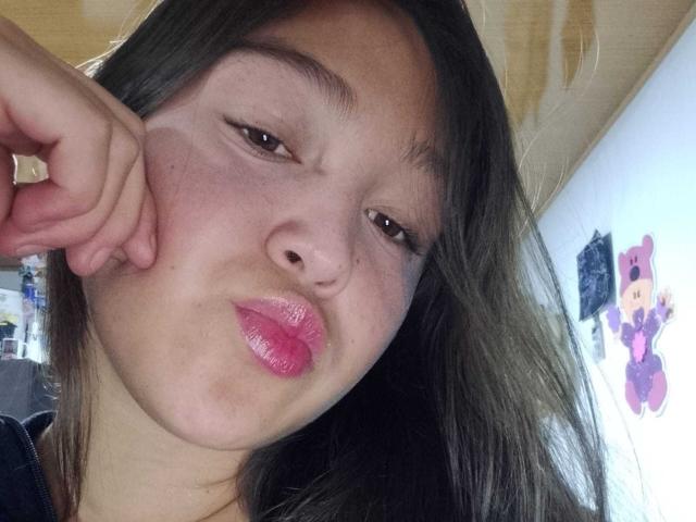 EmmaQuintana's close-up selfie shows a young woman with fair skin, dark brown hair, and pink glossy lips. She has her right hand resting on her cheek, her brown eyes slightly squinted, and a subtle, playful pout. Her hair is straight and falls to the right. The background is blurred, featuring a white wall with a pink teddy bear illustration. The lighting is natural, highlighting her smooth skin and slightly flushed cheeks. The image has a casual, intimate feel.