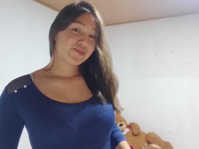 EmmaQuintana with long brown hair, wearing a blue long-sleeve top, smiling slightly, standing in front of a brown teddy bear. Her tits are visible under the tight top.