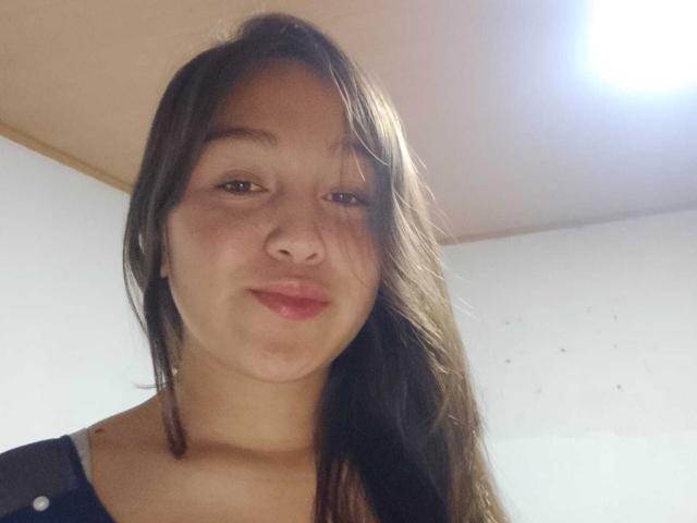 EmmaQuintana with long brown hair, slight smile, and pink lips, looking at the camera, showing her soft brown tits and freckles, in a simple indoor setting.