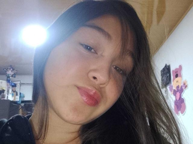 EmmaQuintana with shiny pink lips, slightly flushed cheeks, and dark brown hair in a selfie, eyes half-open, small black nose, light skin, and a subtle smile.