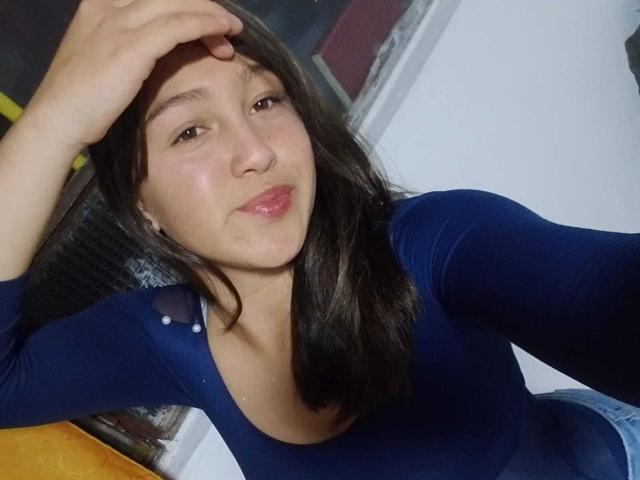EmmaQuintana with brown hair, blue long-sleeve top, pouty lips, and a hand in her hair, showing a slight smile, with visible acne on her cheek.