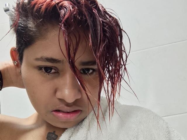 AndromedaSix with wet red hair, pouty face, and white towel, holding her hair up, showing small tattoo on her neck, wearing ear studs, in a bathroom.