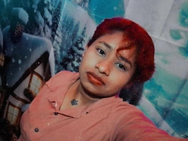 AndromedaSix has short red hair, a medium brown skin tone, and wears a pink button-up shirt. A small heart tattoo is visible on her chest. She looks slightly to the side with a neutral expression. Snowy, mountainous scenery and a small house with a lit window are in the background. The image has a slightly grainy texture and a cool, blue-green tint. Her red lipstick contrasts with her pink shirt. AndromedaSix's right shoulder is closer to the camera.