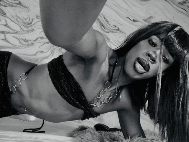 EbonyHansNight with dark, glossy lips, tongue out, wearing a sparkly black bikini top, chain necklace, and short hair, taking a selfie, arms extended, lying on a furry surface.