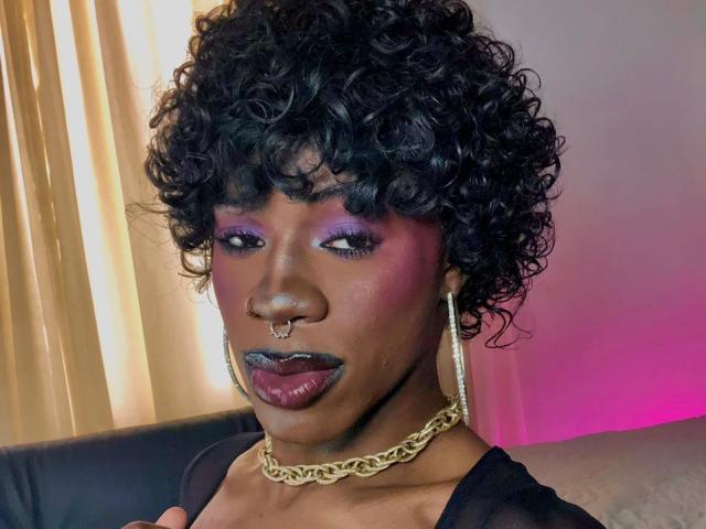 EbonyHansNight has dark curly hair, purple eyeshadow, black lipstick, a gold chain necklace, and a gold nose ring. She's wearing a black top and gold hoop earrings.