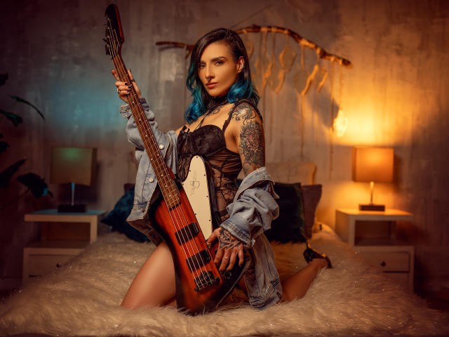EmaLeBlanc with blue hair, tattoos, holding a sunburst bass guitar, wearing black lace lingerie and an unbuttoned denim jacket, kneeling on a fluffy bed.