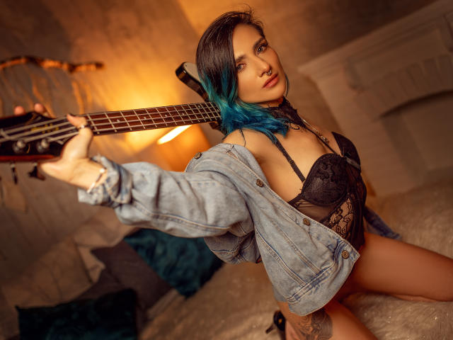 EmaLeBlanc with blue-dyed hair, holding a guitar, wearing a black lace bra, denim jacket, and black panties, exposing her tits and a tattoo on her right thigh.