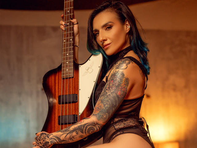 EmaLeBlanc holds a sunburst electric guitar with a white pickguard. She has black hair with blue highlights, a nose ring, and tattoos covering her right arm. She wears a black lace bodysuit, looking at the camera with a slight smile. The guitar's warm wood contrasts with her dark clothing. The lighting is warm, highlighting her tattooed arm and face. Her confident, tattooed appearance dominates the image.