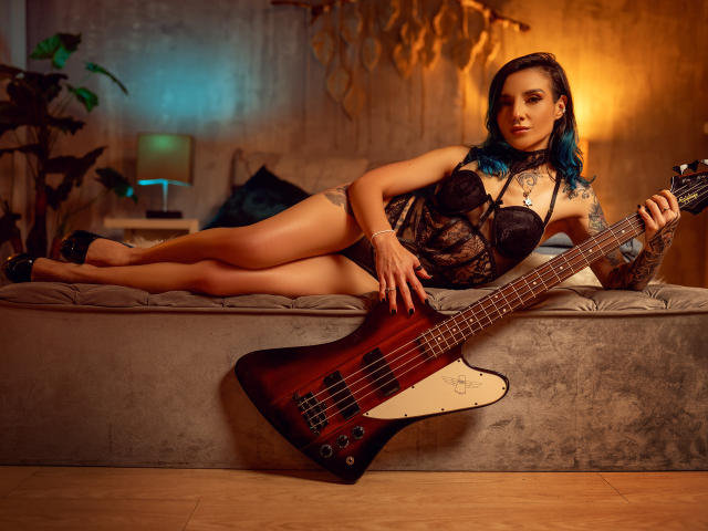 EmaLeBlanc lies on a gray, tufted bench, legs extended, wearing a black lace bodysuit. She holds a red and black electric guitar with a white pickguard. Her blue-dyed hair is loose, tattoos visible on her right arm and chest. Warm, orange light casts shadows on the textured wall behind her. Her black heels and confident posture highlight her toned legs and lace bodysuit. The image is a photograph with a focus on EmaLeBlanc's relaxed yet assertive presence.