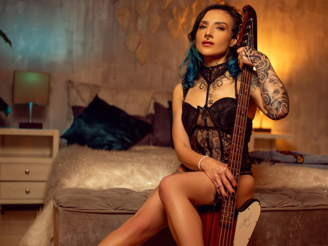 EmaLeBlanc with blue hair, black lace lingerie, tattoos, holding a bass guitar, sitting on a bed, legs crossed, sexy, confident, warm lighting, bedroom setting, sultry expression.