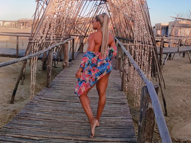 AlexXan with toned ass, bare boobs, long blonde ponytail, holding colorful floral dress, standing on wooden beach bridge, looking back, sand and rustic bridge in foreground.