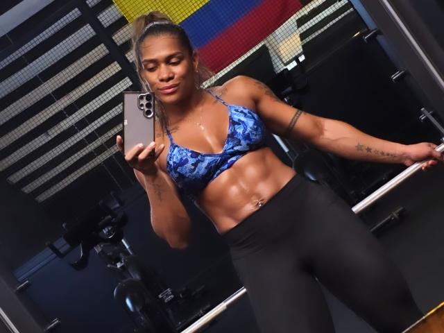 AlexXan, muscular, brown-skinned, blue patterned bra, black leggings, abs visible, arm tattoos, ponytail, holding iPhone, mirror selfie, gym, confident, sexy.