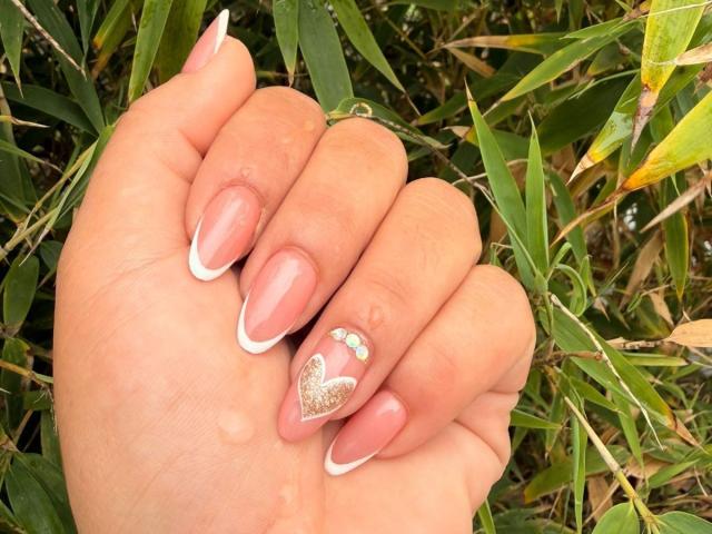ScarlettOxford's hand with pink-nail French manicure, glitter heart on pinky, small green and yellow dots, holding bamboo leaves. Her nails are long and neatly shaped.
