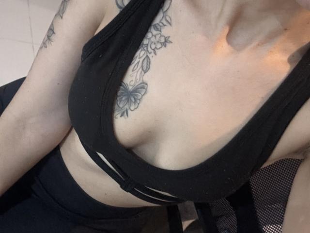 Close-up of LaurentColin's upper body. LaurentColin has a light skin tone and is wearing a black sports bra with a deep V-neck, exposing part of their chest. A detailed black floral tattoo with a butterfly is visible on the left side of their chest and upper arm. LaurentColin's arm is slightly raised, showing more of the tattoo. The texture of the sports bra is smooth, contrasting with the tattoo's intricate lines. The image is slightly angled, focusing on the chest and tattoo. The lighting casts a soft orange glow on the right side of LaurentColin's chest.