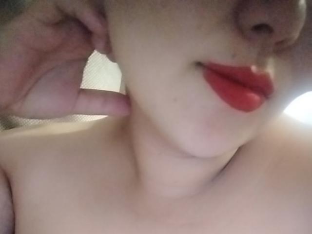 SweeetCheeks' face, showing pale skin, red lipstick, and a hand touching their ear. Boobs and collarbone visible. Soft focus.