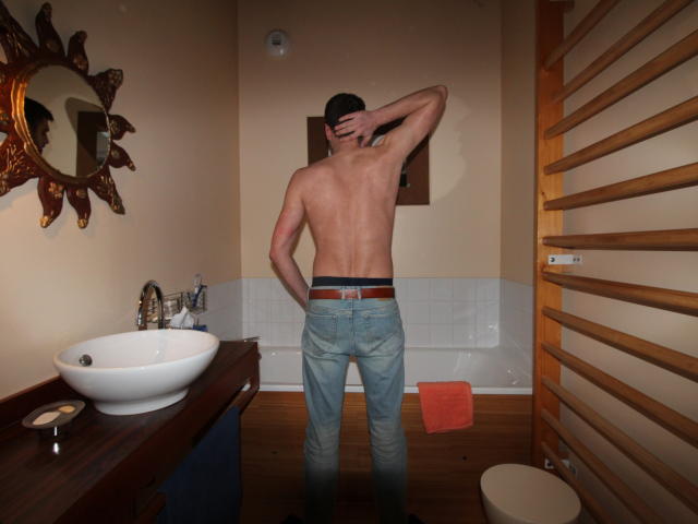 A photograph of Paaul69 standing in a bathroom. He's shirtless, facing away, with one arm raised, hand touching the back of his head. He's wearing blue jeans with a brown belt. The room has a wooden towel rack on the right, a white bathroom sink on the left with a round mirror and a sun-shaped decorative wall piece above it. A white tile backsplash and a red towel are visible. The floor is wooden.