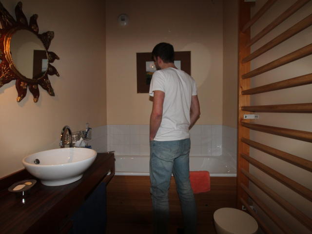 Paaul6 stands in a small bathroom with his back to the viewer. He wears a white t-shirt and blue jeans. The room has a wooden vanity with a white bowl sink on the left, a sun-shaped mirror above it, and a towel on the floor. He faces a white-tiled wall with a wooden radiator on the right and a toilet below it. The floor is wooden. The image has a casual, realistic style.