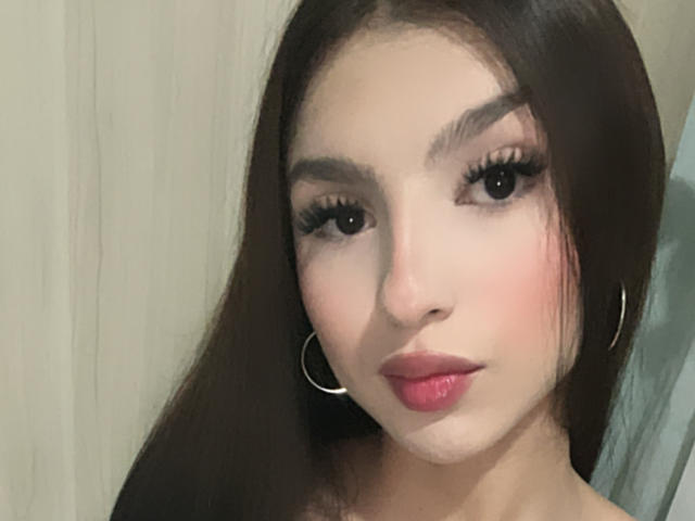MilanMoon has long brown hair, thick eyebrows, long eyelashes, pink lipstick, and wears silver hoop earrings. She has a light skin tone and a slightly flushed face.