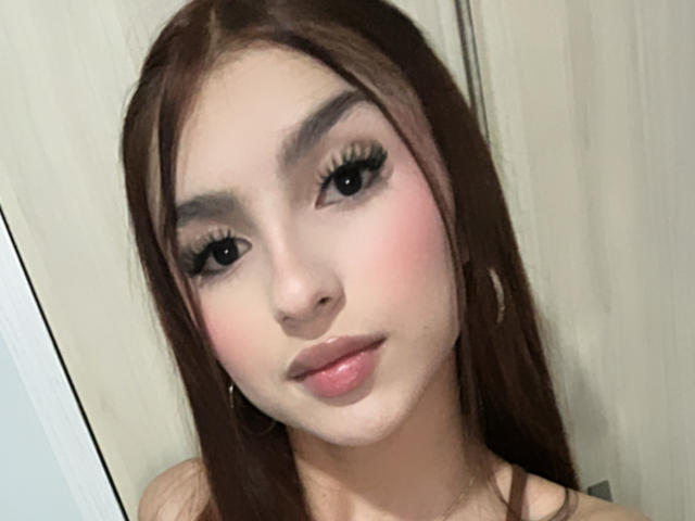 MilanMoon has long brown hair, thick eyelashes, pink blush, and glossy pink lips. She's wearing hoop earrings and looking at the camera with a slight smile.