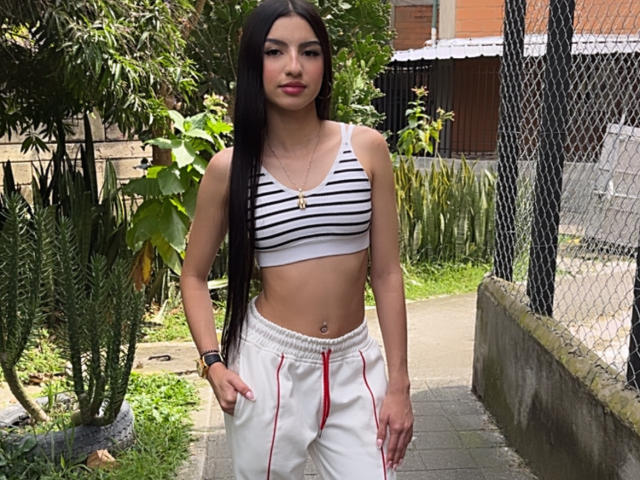 MilanMoon has long black hair, small tits, wearing a striped crop top and white pants with red stripes, standing outdoors, hand in pocket, looking at camera.