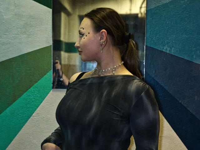 AriaVerlangen-hot with dark brown hair in a ponytail, wearing a black leather off-shoulder top, pearl necklace, and black earrings, standing against a green and blue striped wall.