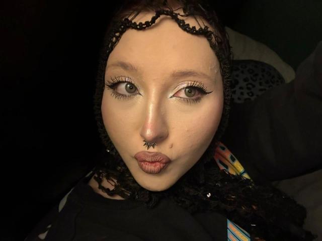 Close-up of AriaVerlangen-hot with light skin, green eyes, and black lace headpiece. She has a black star-shaped nose ring and puckered lips. She's wearing thick, black eyeliner with a slight wing and long lashes. Her hair is partially visible, curly, and black. She's dressed in a dark top with colorful patterns. The lighting is dark, focusing on her face. Her expression is playful and slightly provocative. The texture of her lace headpiece contrasts with her smooth skin.
