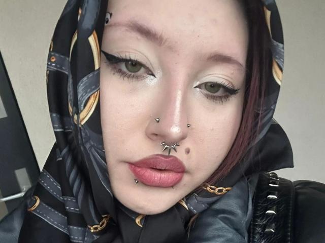 AriaVerlangen-hot with pale skin, green eyes, black hood, facial piercings (nose, septum, lip), red lipstick, and dark makeup. She has a red side-swept hair and a black jacket.