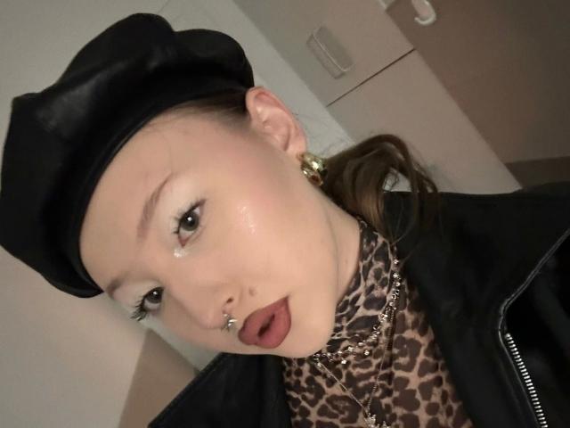 AriaVerlangen-hot with a black leather cap, leopard print shirt, gold earrings, nose ring, and lip ring, looking surprised with her mouth open. Sheer skin, brown eyes, and brown hair.