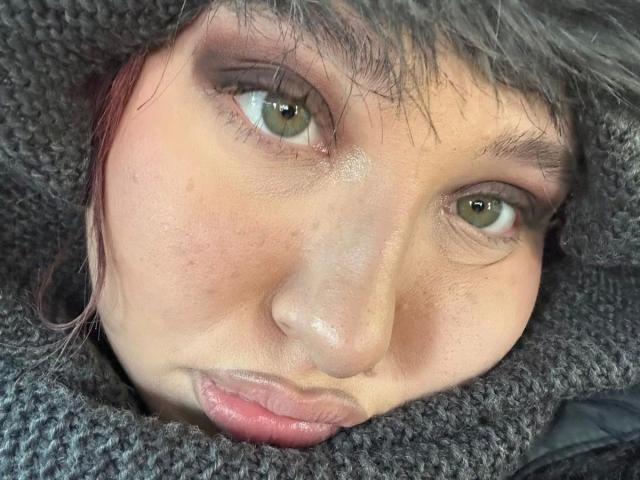 Close-up photo of AriaVerlangen-hot's face, eyes looking slightly upward. AriaVerlangen-hot has light brown skin, green eyes, and subtle pinkish blush on the cheeks. Wet, dark brown hair peeks out from a gray, textured wool hat that covers most of her head. AriaVerlangen-hot's lips are slightly parted, showing a hint of pink. The texture of the wool hat is rough, contrasting with her smooth skin. Soft light highlights her eye's greenish-brown color. AriaVerlangen-hot's expression is neutral, almost pouting.